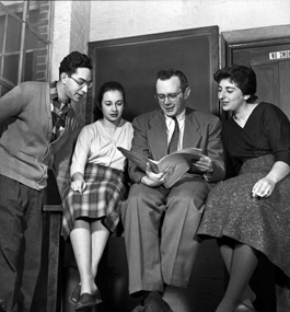 Professor Irving Howe, reading a book, with three of his Brandeis students gathered around him.